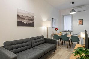Een moderne woonkamer met een grijze bank, een eethoek met tafel en stoelen, en een keuken met een raam.