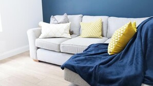 Ein hellgraues Sofa mit gemusterten Kissen vor einer dunkelblauen Wand, eine blaue Decke auf dem Sofa und ein heller Holzboden.