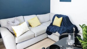 Ein modernes Wohnzimmer mit einem grauen Sofa, gelben Kissen mit Muster, einer blauen Decke sowie einem Essbereich mit einem Glastisch und Stühlen.