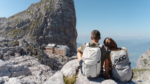 Duas pessoas com mochilas numa montanha rochosa, com vista para um edifício com várias janelas.