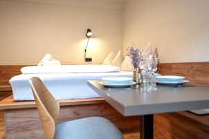 Una habitación moderna con cama, mesita de noche con lámpara y zona de comedor con mesa puesta para dos.