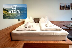 Ein Doppelbett mit weißen Leinen und Handtüchern, ein hölzernes Kopfende, ein an der Wand befestigtes Fernsehgerät, das 'HOCHKÖNIG CARD' anzeigt, sowie gerahmte Bilder an der Wand.