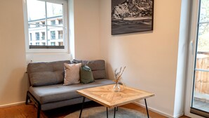 Sala de estar com um sofá cinzento, mesa de centro de madeira, tapete, janela e um quadro na parede.