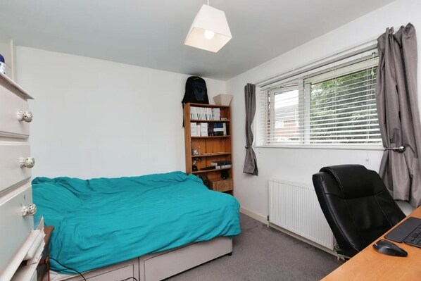 A bedroom with a bed, a desk with a chair, a bookshelf, and a window with blinds.