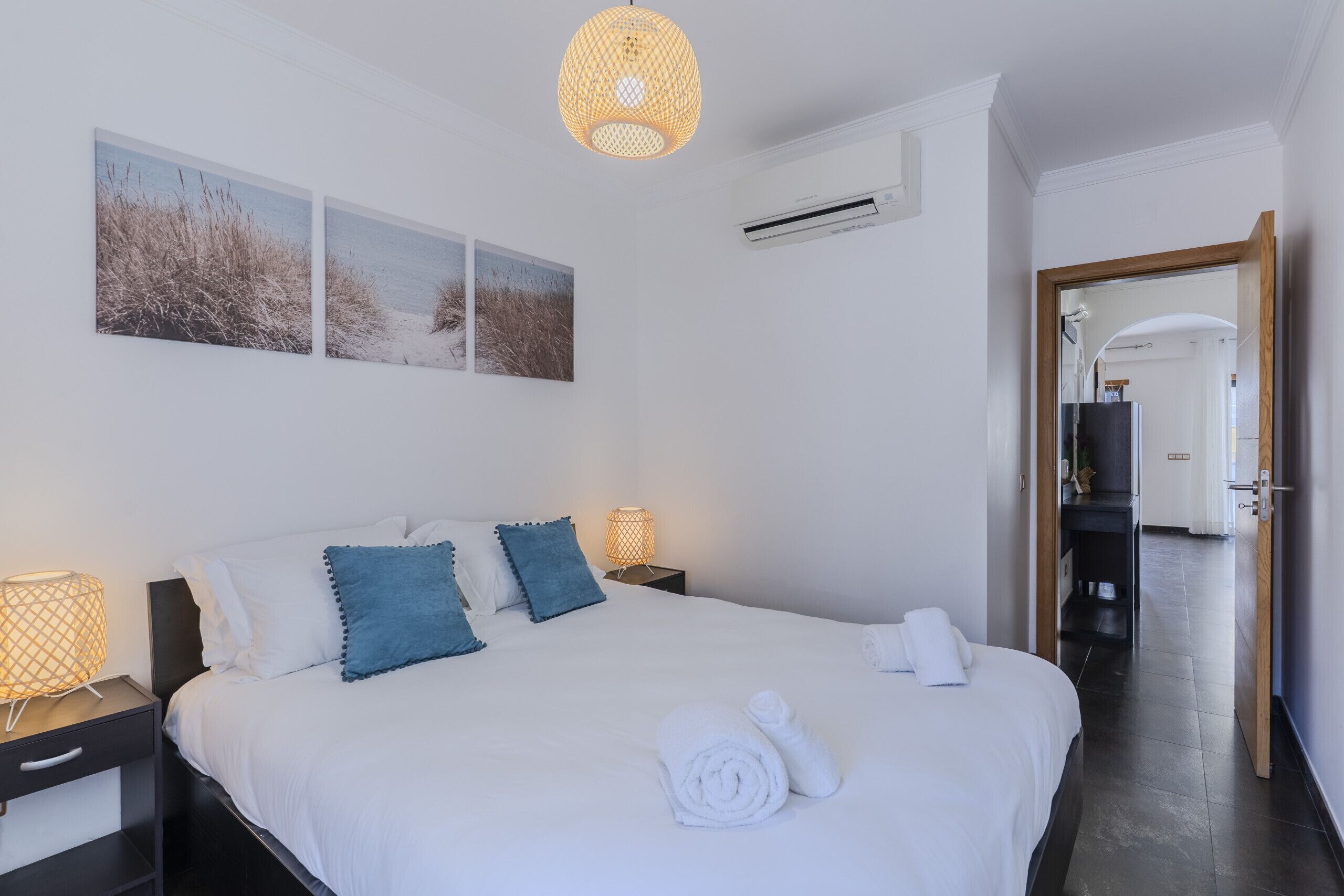 Habitación de hotel con cama, lámparas de noche, aire acondicionado instalado en la pared y un pasillo que lleva a un baño.