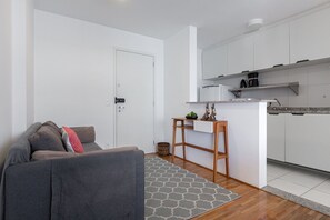Ein modernes Apartment mit grauem Sofa, einem kleinen Holztisch und einer Küchenzeile mit weißen Schränken und einer Kaffeemaschine.