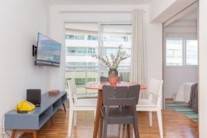 Ein modernes Hotelzimmer mit Balkon, einem an der Wand befestigten Fernseher, einem kleinen Tisch mit Stühlen und einer Vase mit Blumen auf dem Tisch.
