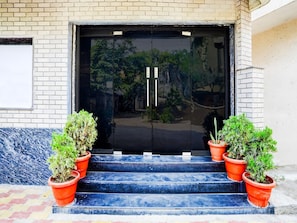 A building entrance with a black door, two potted plants on the steps, and a brick wall.