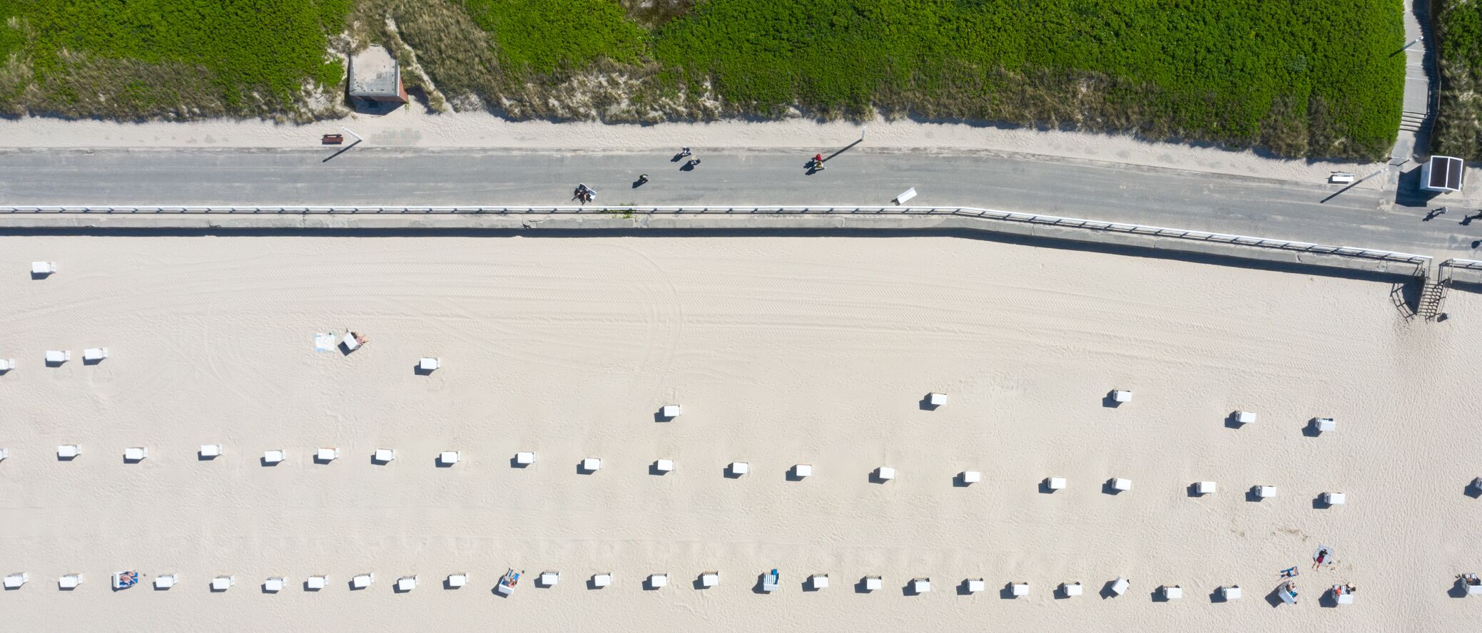 Luchtbeeld van een strand met rijen witte strandhutten en een verharde promenade langs de kust.