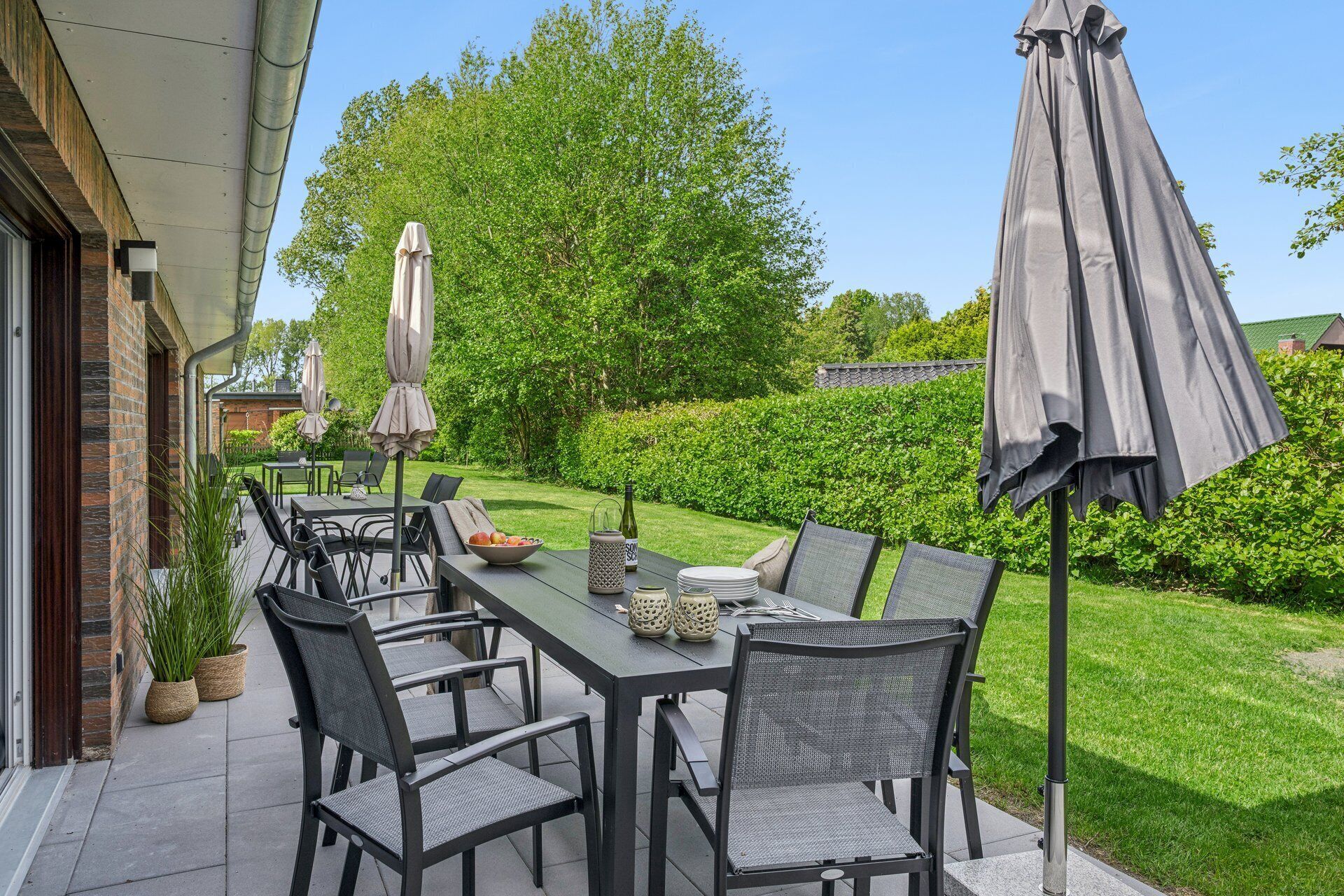Eine Terrasse im Freien mit Tisch und Stühlen, umgeben von Pflanzen und einer Hecke.