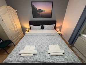 Ein Schlafzimmer mit einem gemusterten Bettbezug, zwei Kissen, einem Kopfteil, Nachttischlampen, einem Nachttisch, einem Kleiderschrank und einem gerahmten Bild an der Wand.