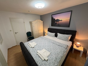 Ein Schlafzimmer mit Bett, Kleiderschrank, Sessel, Nachttisch mit Lampe und einem Bild an der Wand.