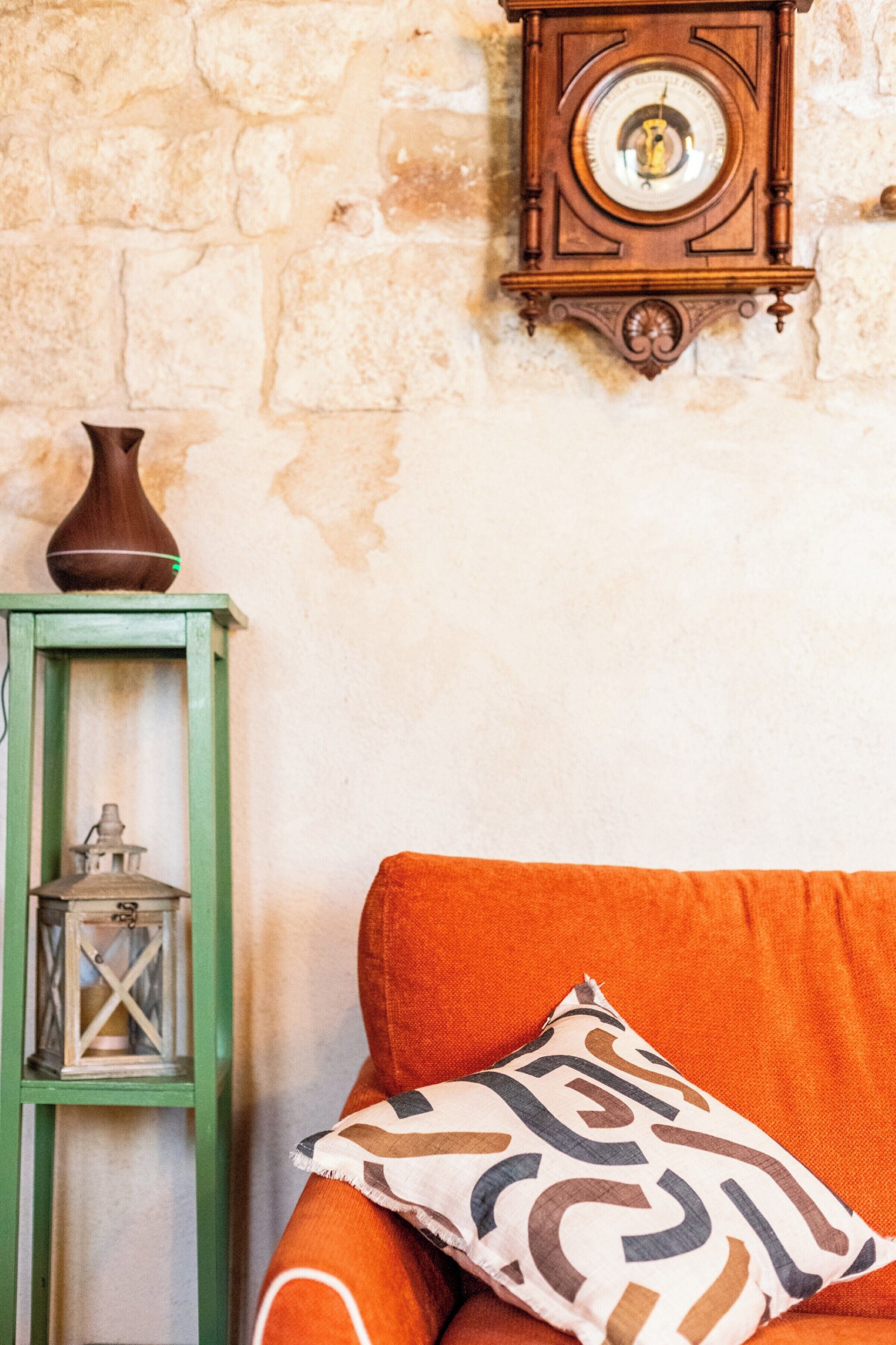 A room with a stone wall, a green side table with a lantern, and an orange sofa with patterned pillows.