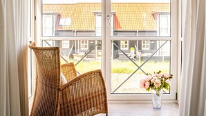A room with a wicker chair, a vase with flowers, and a view of a building with a tiled roof. - 5 Star Holiday Home in Juelsminde (Juelsminde)