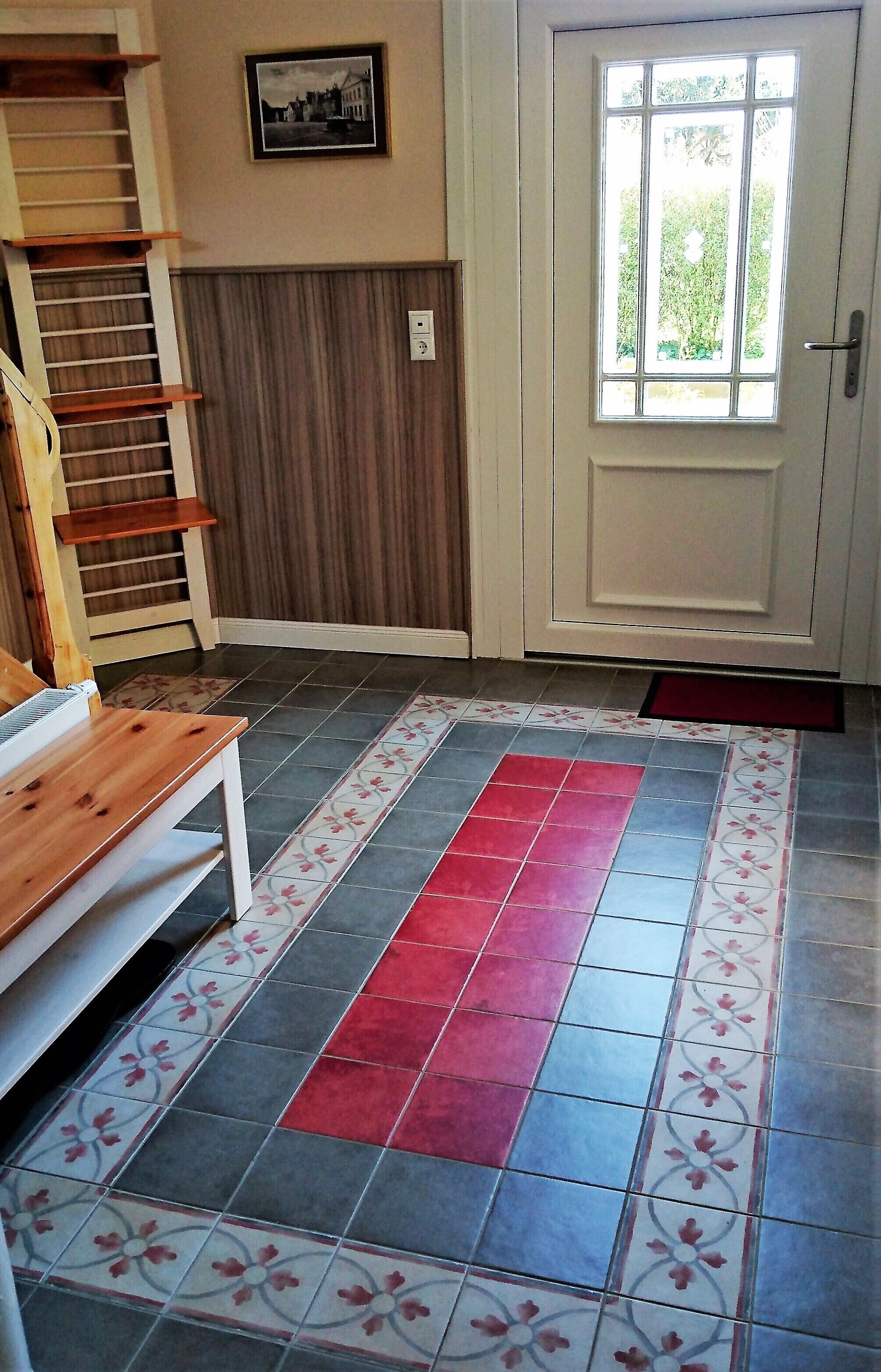 A room with a wooden bench, a tiled floor with a red strip, a door with a window, and a wall-mounted picture.