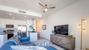 A modern living room with a sofa, ottoman, TV, and a kitchenette in the background. - Modern Bayfront Condo with Pool & Balcony View (Sarasota)