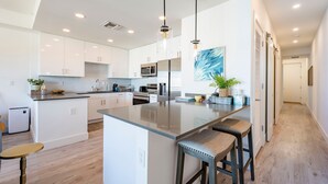 A modern kitchen with white cabinets, a central island, pendant lighting, and a view into the adjacent living area. - Modern Bayfront Condo with Pool & Balcony View (Sarasota)