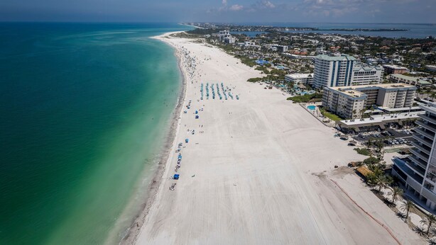 An aerial view of a beach with a long stretch of sand, a few umbrellas, and buildings adjacent to the shore. - Modern Bayfront Condo with Pool & Balcony View (Sarasota)
