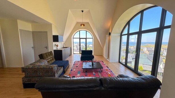 A spacious loft with a high ceiling, large windows offering a city view, a black leather sofa, and a patterned rug. - Avel Vor - Family home, renovated, sea view (Porspoder)