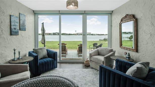 Ett rum med en stor skjutdörr i glas som leder ut till en balkong med utsikt över havet och en avlägsen stadsbild. Inuti finns det två blå soffor, en beige fåtölj och en mönstrad pall.