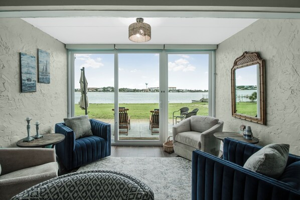A room with a large sliding glass door leading to a balcony with a view of the sea and a distant cityscape. Inside, there are two blue sofas, a beige armchair, and a patterned ottoman. - Sea Palm View 5E (Fort Walton Beach)