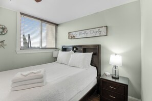 A neatly made bed with white linens, a bedside table with a lamp, and a wall decoration. - Sea Palm View 5E (Fort Walton Beach)