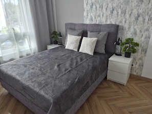 A modern bedroom with a grey patterned bedspread, a grey upholstered headboard, two bedside tables with lamps, and a small potted plant on each. - Horizon B9 by Grand Apartments (Gdańsk)