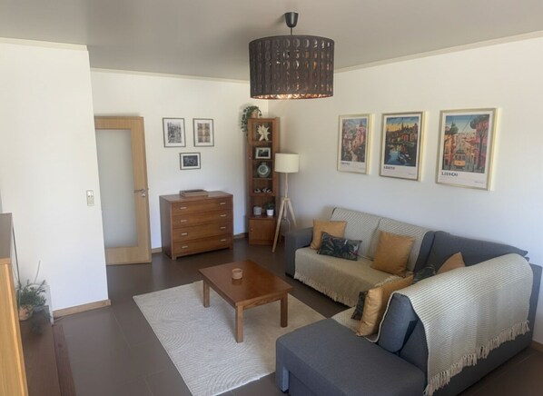 Una sala de estar con sofá, mesa de centro y lámpara. Hay cuadros en la pared y un mueble de madera.