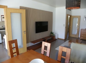 A modern living room with a flat-screen TV mounted on a wall, wooden furniture, and a dining table.