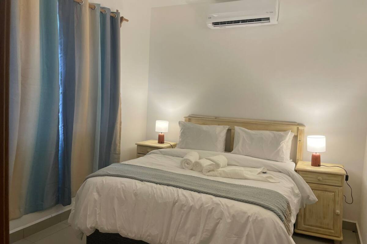 A bedroom with a bed, two bedside tables with lamps, a wall-mounted air conditioner, and a curtain.