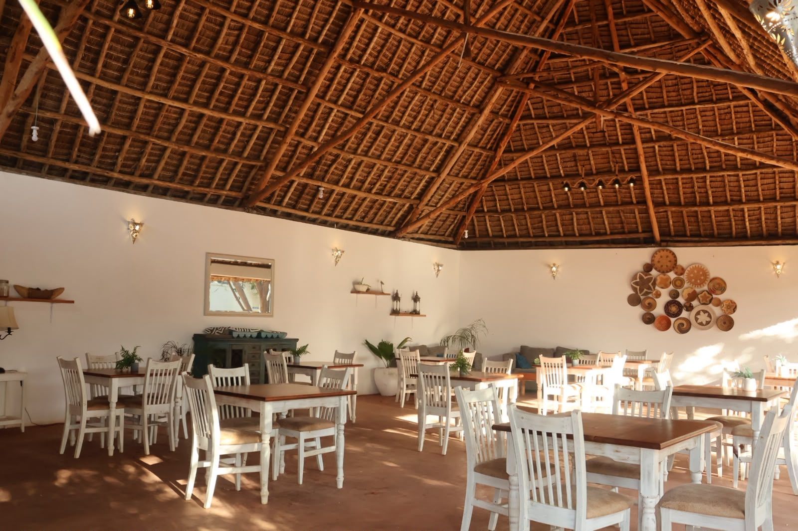 An open-plan dining area with wooden tables and chairs, a thatched roof, and wall-mounted light fixtures.