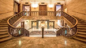 A grand staircase with ornate railings, a marble floor, and a patterned carpet leading to a room with a large wooden door and a chandelier.