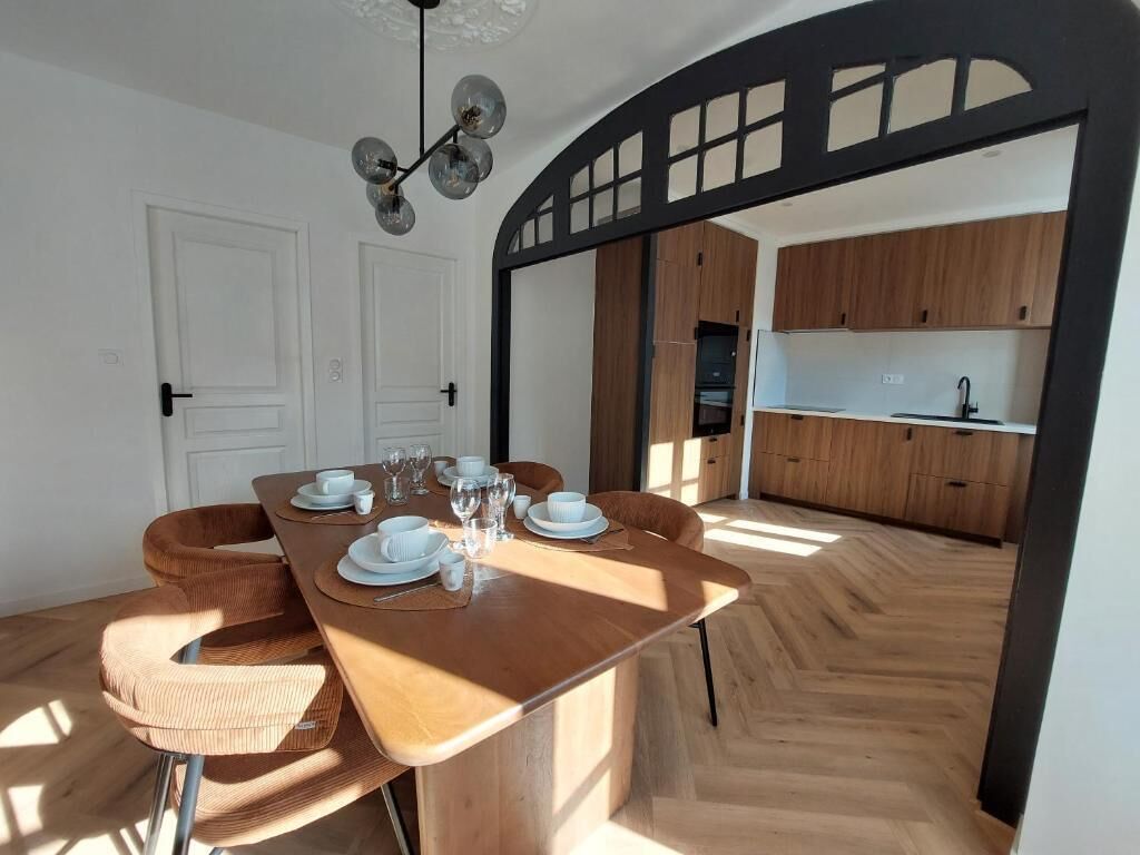 Ein Essbereich mit einem Holztisch, gedeckt für vier Personen, eine durch einen Bogen sichtbare Küche und eine Deckenleuchte.