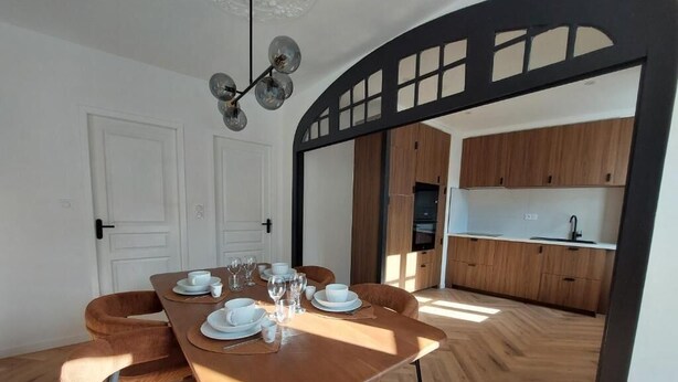 A dining area with a wooden table set for four, a kitchen visible through an archway, and a ceiling light fixture.