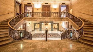 A grand staircase with ornate railings, a marble floor, and a patterned carpet leading to a room with a large wooden door and a chandelier.