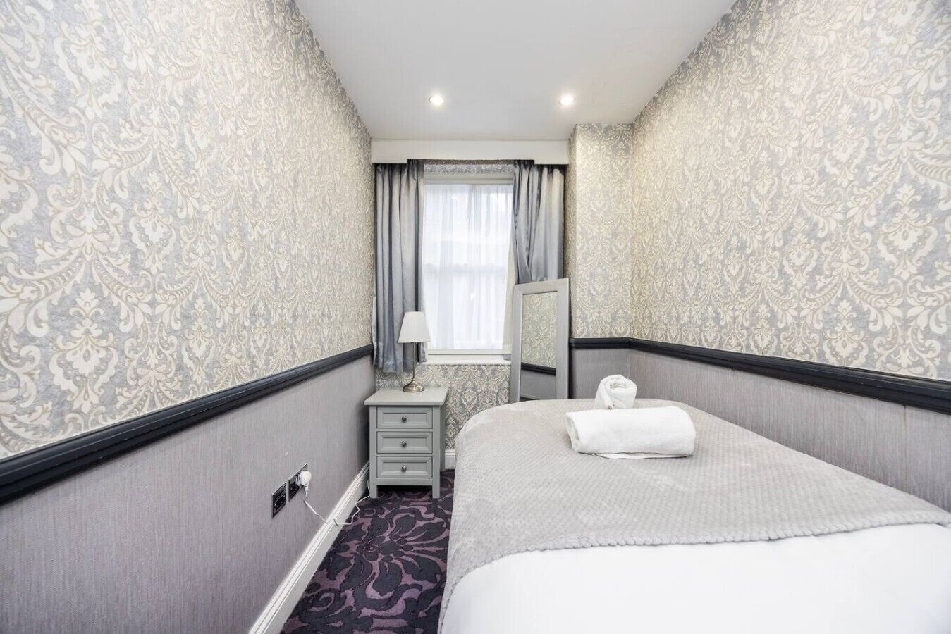 Une chambre d’hôtel avec un lit, une table de chevet, une lampe, un miroir et une fenêtre avec des rideaux.