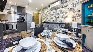 Une cuisine moderne équipée d’appareils électroménagers en acier inoxydable, un coin repas avec une table ronde pour quatre personnes et un salon avec un canapé et un mur orné d’un motif bleu.