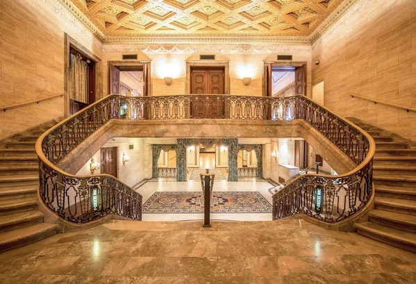A grand staircase with ornate railings, a marble floor, and a patterned carpet leading to a room with a large wooden door and a chandelier.