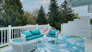 A covered outdoor deck with white railings, two white wicker sofas with teal cushions, and a patterned floor mat.
