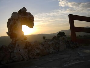 Eine Steinstruktur mit einem natürlichen Torbogen, einem Holzrahmen und Blick auf einen Hügelland untergehendem Sonnenuntergang.