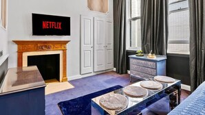 Une pièce avec une cheminée, un téléviseur affichant le logo « NETFLIX », une commode, une fenêtre avec des rideaux et un tapis bleu.