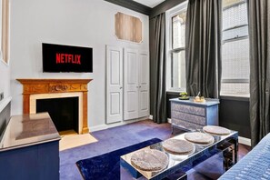 Ein Zimmer mit Kamin, einem Fernseher, der 'NETFLIX' anzeigt, einem Schminktisch, einem Fenster mit Vorhängen und einem blauen Teppich.