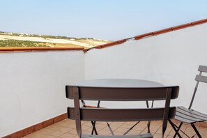 Un pequeño balcón al aire libre con una mesa y sillas, una pared blanca y vistas a un paisaje distante.