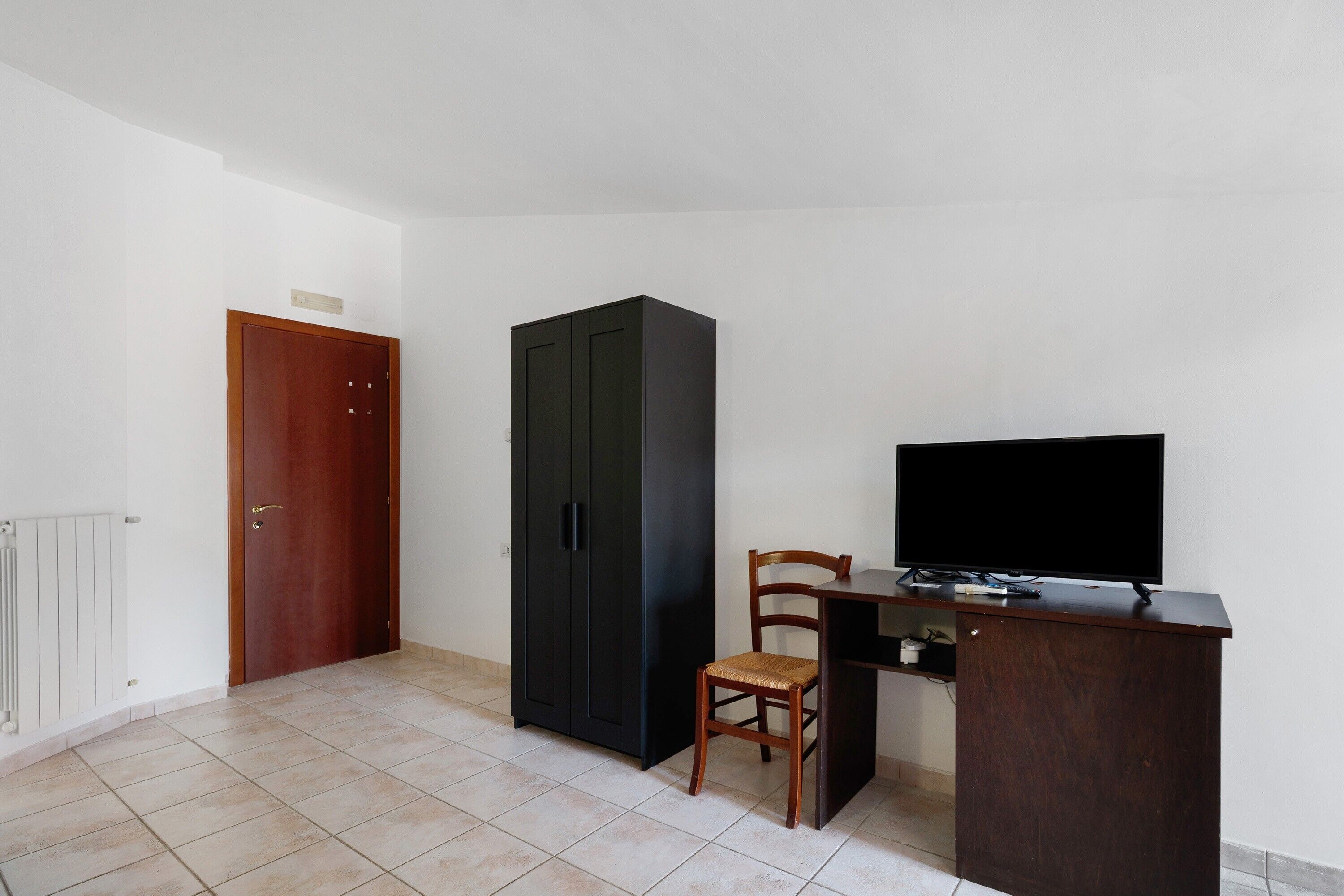 Una habitación con puerta de madera, un armario negro, una silla de madera, un televisor sobre un mueble de madera y suelo de baldosas.