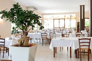 A restaurant with a large potted tree in the foreground, multiple tables with white tablecloths, and a bright interior. - Villa Ristòries Room 6 with Terrace, Wi-Fi and Air Conditioning (Matera)