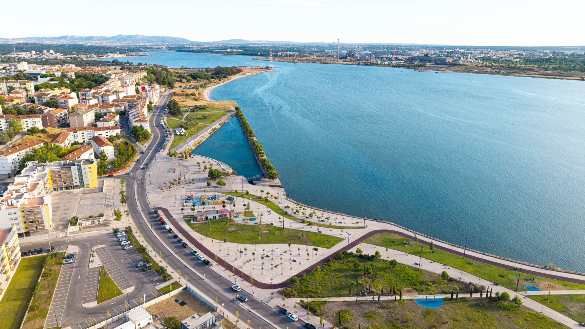 An aerial view of a coastal city with a marina, a curved road, and a mix of residential and commercial buildings.