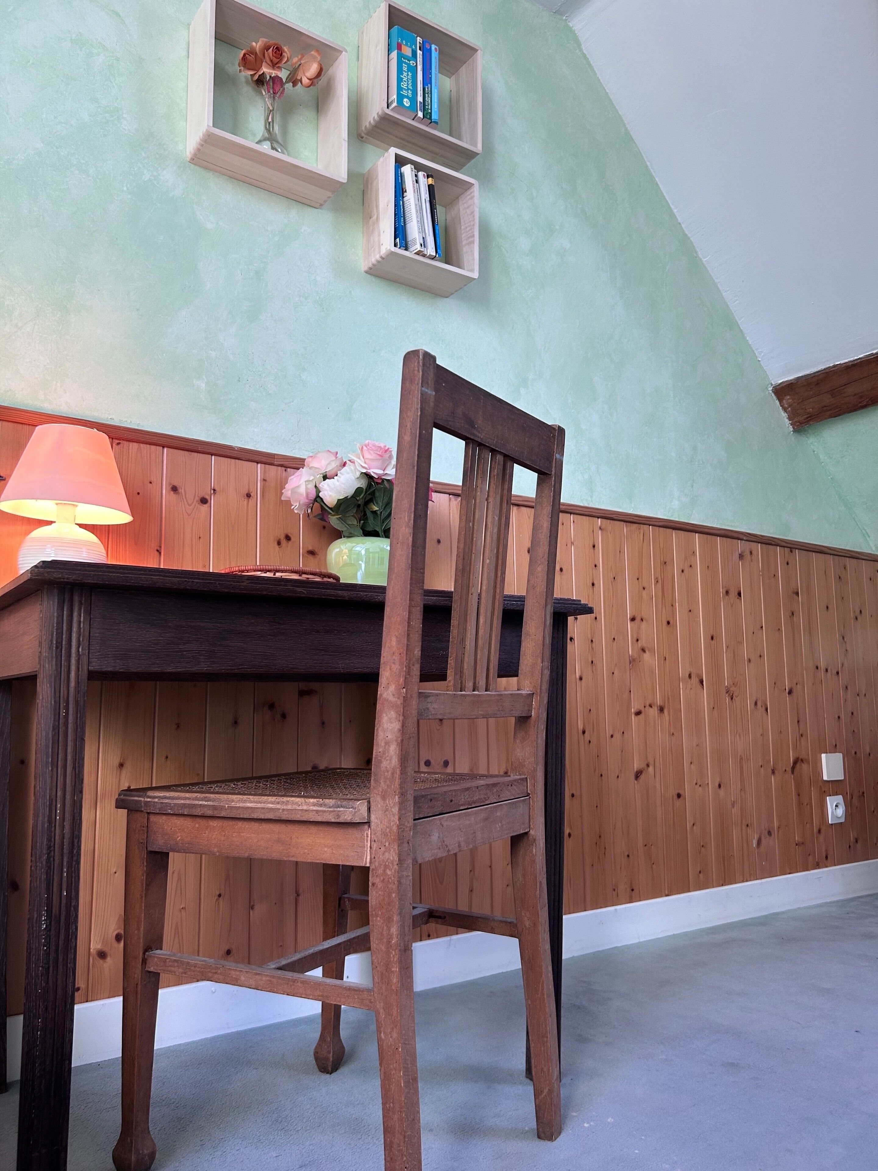 Una silla de madera frente a una mesa con una lámpara y un florero. Hay estantes de madera sobre la mesa con libros y otro florero.
