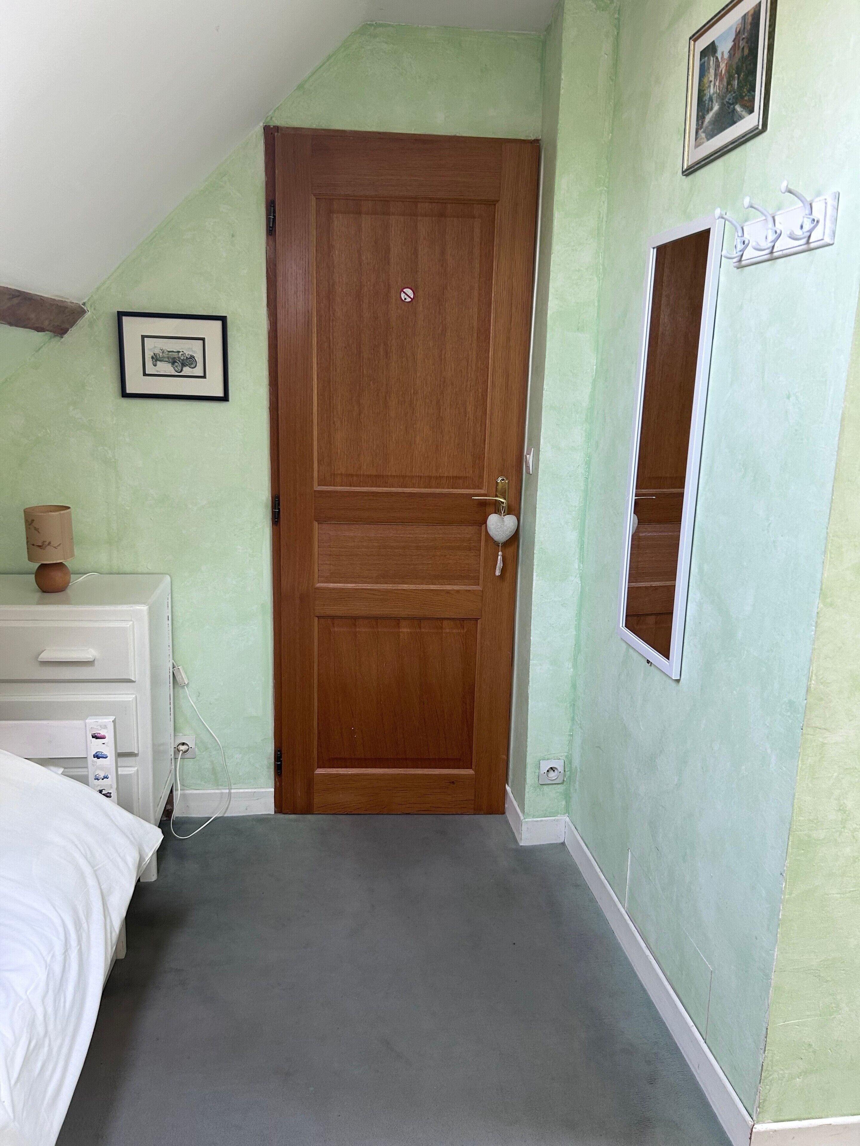A room with a wooden door, a bedside table with a lamp, a dresser, and a small window.