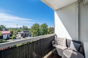 Ein Balkon mit einer Rattan-Couch und einem kleinen Tisch, mit Blick auf eine Wohngegend mit Bäumen und Häusern.