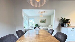 A dining area with a wooden table, four chairs, a chandelier, and a view into a living room with a sofa and a painting. - Modern and family friendly house in Reykjavik (Reykjavík)